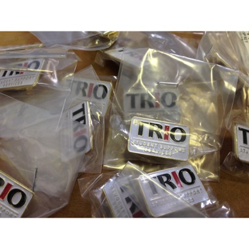 TRIO Student Support Services Lapel pin. Sold as shown. Bag of 50 pins. "TRO" capital letters in black, with "I" in red. 50 lapel pins per order. 1" x ~ 1/2" Die Struck Lapel Pin Bright Goldtone Finish Black, White, Red (PMS 186c) Soft Enamel Colorfill. Individually polybagged